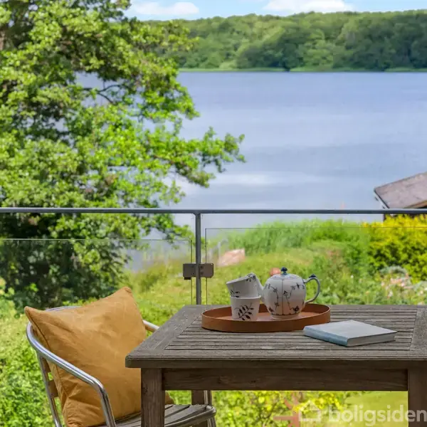 En træbord står med en kaffekande og kopper på en terrasse. Det er omgivet af grønne træer og har udsigt over en sø og skov.