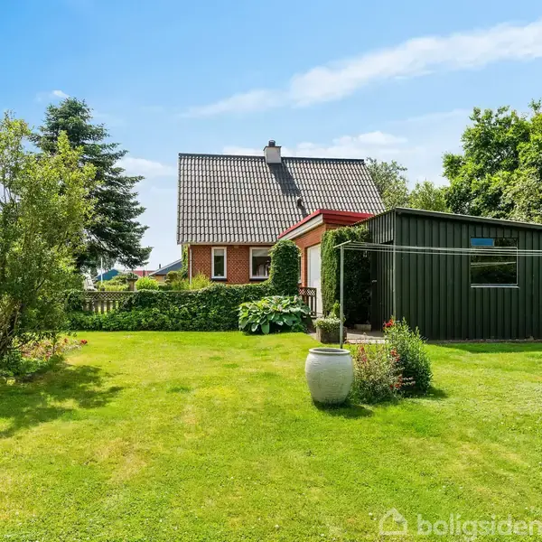Rødt hus med sort tag står bag en grøn have med græsplæne og buske. Til højre er en mørkegrøn skurbygning. Sollys belyser området under en blå himmel.