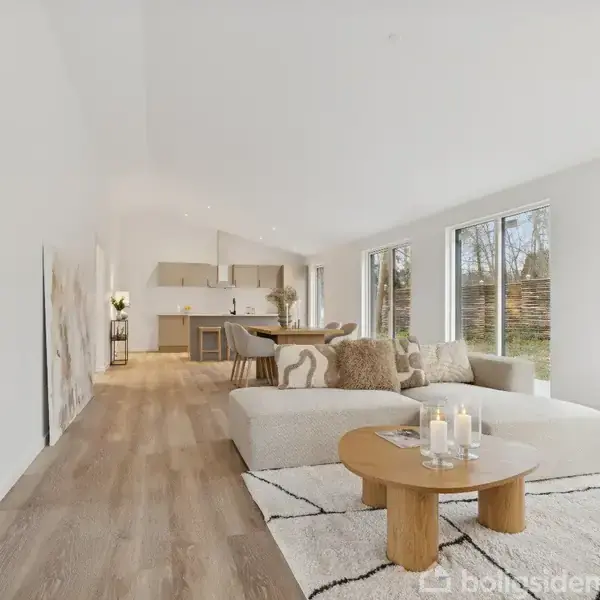 Lys stue med hvid sofa og træbord med tændte stearinlys, placeret på et lyst tæppe. Store vinduer med udsigt til en have i baggrunden.