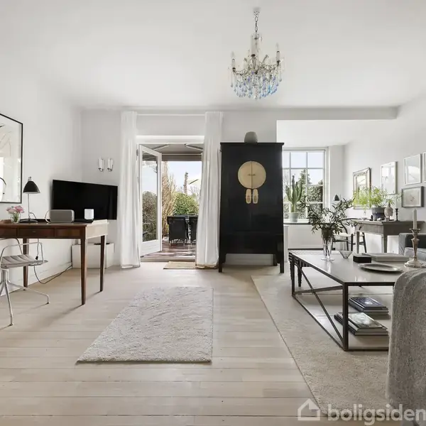 Stue med træbord ved siden af sofa, glasbord foran grå lænestol, og sort reol med ur. Store vinduer og glasdør åbner til terrasse med planter.