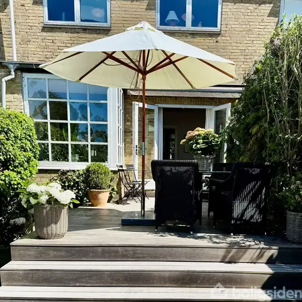 Terrasse med parasol står over siddepladser, omgivet af planter i krukker. I baggrunden ses en bygning med store vinduer og åben dør, der fører ind til husets interiør.