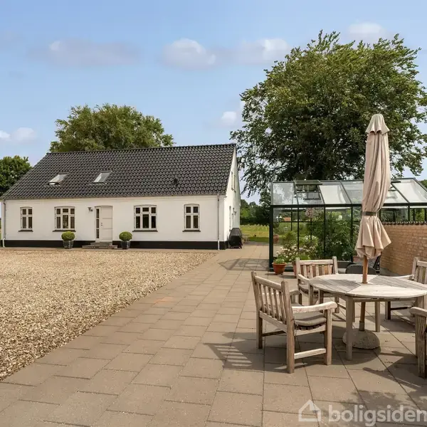 Hvidt hus med skråtag står i baggrunden. Foran er der en terrasse med runde havemøbler og parasol. I nærheden ses et drivhus, grusgård og grønne træer i marklandskab.