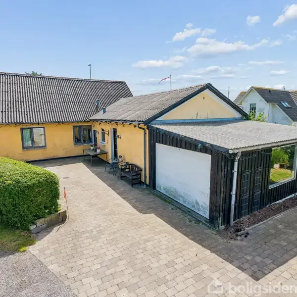 Gult enfamiliehus med mørkt tag har en stor indkørsel og en overdækket terrasse. Huset er omgivet af en velplejet hæk, og der er flere nabohuse i baggrunden under en blå himmel.
