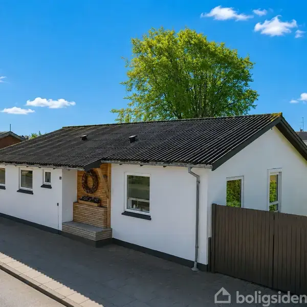 Hvidt hus med sort tag i et villakvarter. Solen skinner, og der er en stor, grøn træ i baggrunden. Huset har en indgangsparti med naturlige trædetaljer.
