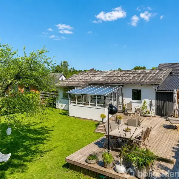 Hus med solterrasse rummer havemøbler på trædæk, omkranset af en frodig grøn have med træer og gyngesofa. Baggrunden viser en klar blå himmel med spredte skyer.