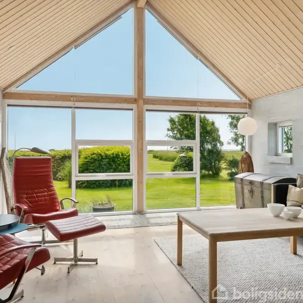 Stue med røde lænestole og sofa står stille foran stort vindue, som viser en solrig have og blå himmel. Lyse trævægge og lofter omgiver rummet med moderne indretning.