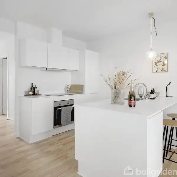 Et moderne køkken med hvide skabe har en central ø med barstole. På øen står flaske, glas og en vase med tørrede blomster. Rummet har lyst trægulv og hvide vægge.