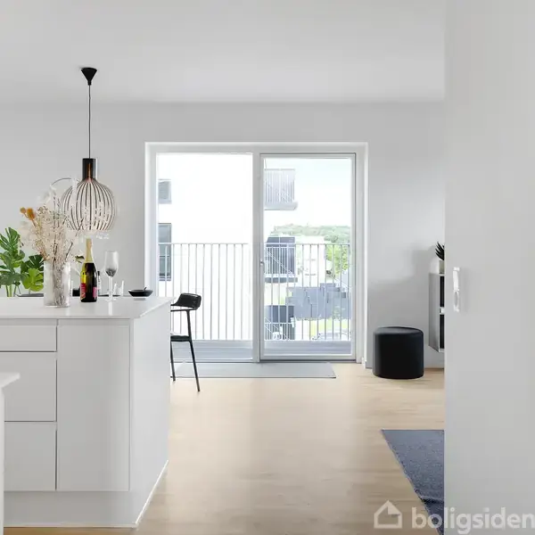 En lys stue med et moderne køkkenbord fyldt med flasker og planter, står til venstre. Store skydedøre fører ud til en altan. Enkel indretning i skandinavisk stil.