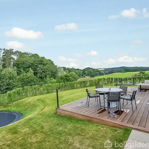 Terrasse med havemøbler på trædæk står ved siden af en moderne bygning. En græsplæne med nedsænket trampolin omkranser området, mens frodige grønne træer og bakker udgør baggrunden.