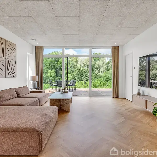 Brun sofa står i en stue med trægulv, hvor en stor fladskærm hænger på væggen. Store vinduer viser en terrasse og frodigt grønt udenfor.