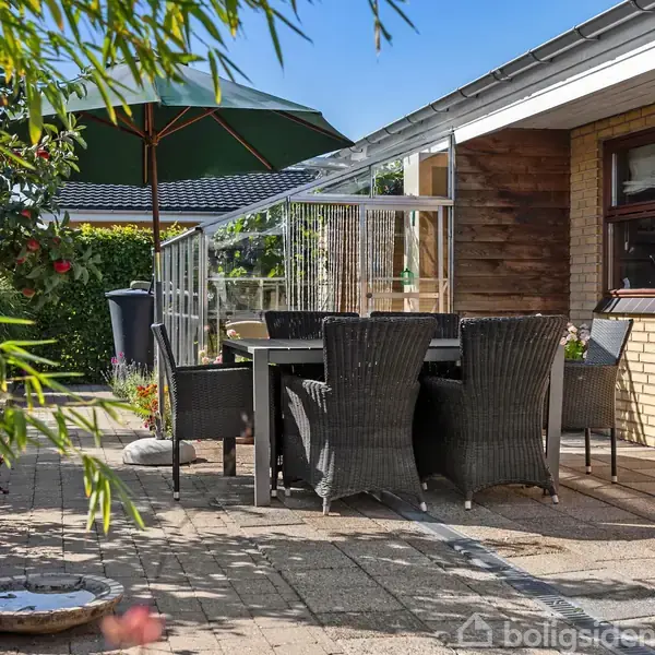 Terrasse med spisearrangement omfatter flettede stole omkring et bord. Solparasol skaber skygge. Murstenshus og grøn beplantning omgiver området med klare, solrige himmel foroven.