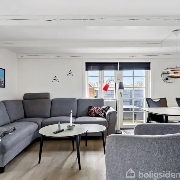 Grå sofa med tæpper står ved langbord i en stue, oplyst af store vinduer. Runde borde og stole omkring sofaen; vægge prydet med kunst. Ingen tekst til stede.