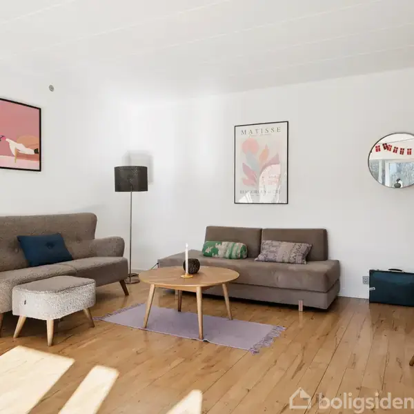 Stue indeholder to sofaer og et sofabord. En stålampe står ved siden af en sofa. Væggen har kunstindramning. Murstensvæg i baggrunden. Trægulv og spejl til højre.