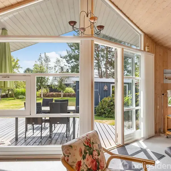 En stue med store vinduer åbner ud til en terrasse med havemøbler og parasol. Indendørs er der lyse trævægge, en lænestol med blomsterprint, og en lædersofa.