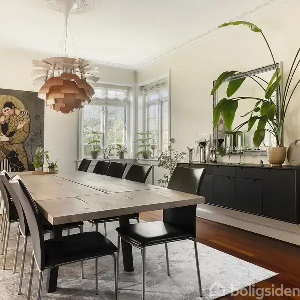 Et langt træbord med sorte stole står i en spisestue. En moderne lysekrone hænger over bordet, og et maleri pryder væggen. Store planter og et skab ses i baggrunden.