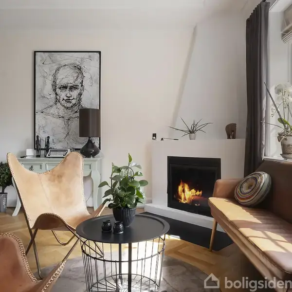 Stue med moderne indretning; ild i pejsen skaber hygge. Et rundt bord med en plante foran læderstole og sofa. Store vinduer slipper naturligt lys ind, malerier på væggene.