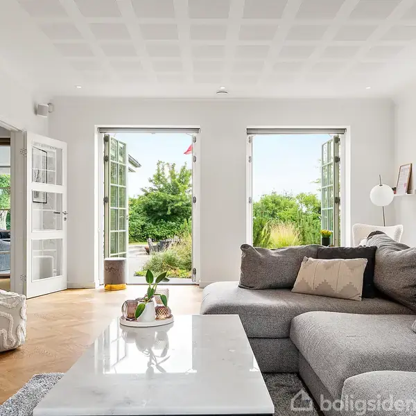 Grå sofa står i en lys stue med hvide vægge. Døre åbner ud til en frodig have, mens et marmor sofabord og en puder dekorerer rummet.