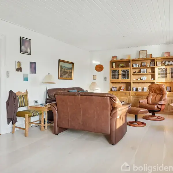 Stue med brune lædersofaer står midt i rummet. En reol med bøger og dekorationer fylder bagvæggen. Lys træmøbler, billeder på væggene, og et vindue til højre skaber en hyggelig atmosfære.