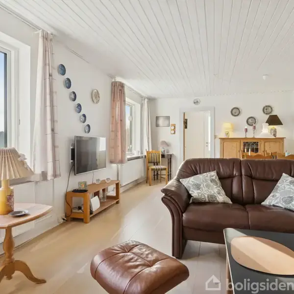 En brun sofa med puder står i et lyst, indrettet stueområde med trægulv. Et lille bord og en lænestol står ved siden af vinduer, som giver udsigt til natur udenfor.