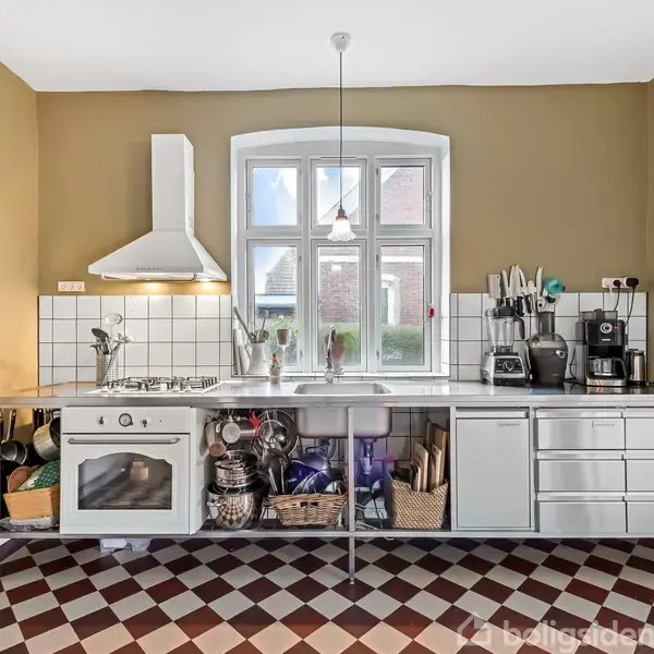 Et moderne køkken med stålbord indeholder en ovn, komfur, kaffemaskine og opbevaring. Rummets vægge er lysebrune, og skakternet gulv dekorerer det. Et vindue tillader naturligt lys.