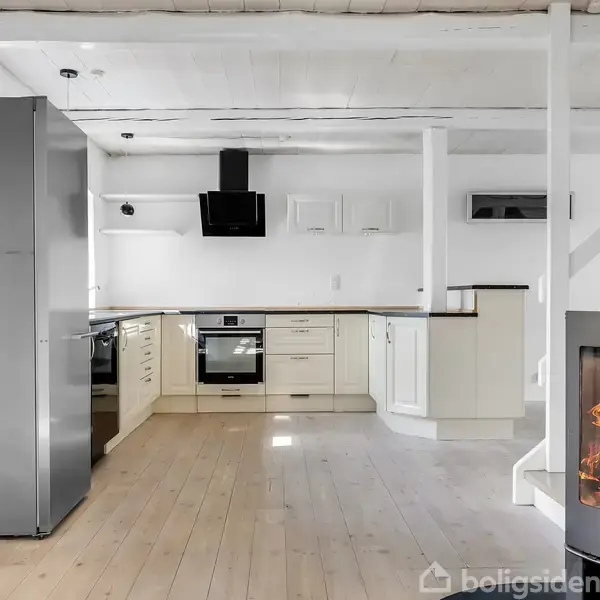 Et moderne køkken med hvide skabe og en sort emhætte. En brændeovn med flammer står til højre på et træparketgulv. Rummet er lyst og åbent.