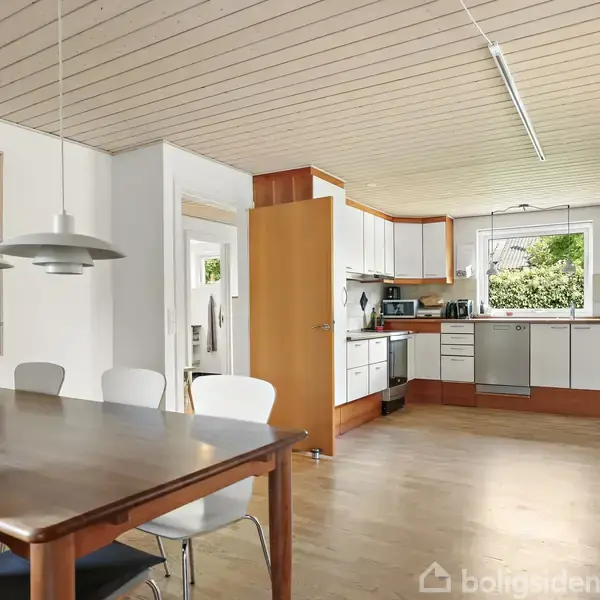 Træbord med stole står i midten af et lyst køkken-alrum. Køkkenet har hvide skabe og vindue med udsigt til have. Lamper hænger over bordet, og der er et kunstværk på væggen.