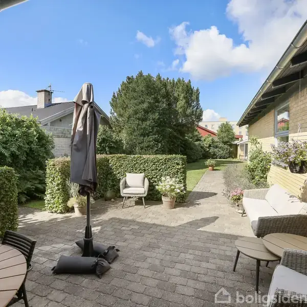 En terrasse med runde borde og stole er beliggende i en solrig have omkranset af buske og træer. En sammenklappet parasol står ved siden af møblerne.