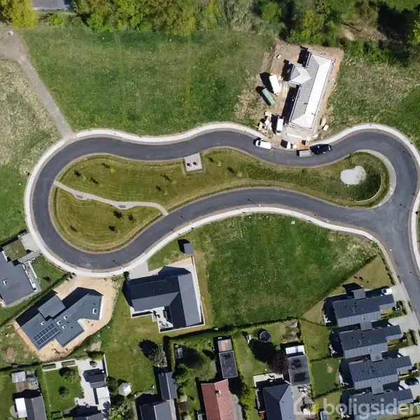 En snoet vej løber gennem et grønt område med et enkelt hus under opførelse. Omkring vejen ses flere boligområder med huse og haver.
