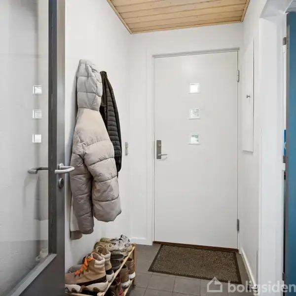 Dør i entre prydet med frakker hængende på kroge; et par hylder med sko langs væggen; lyst træloft og grå flisegulv danner en minimalistisk indgang.