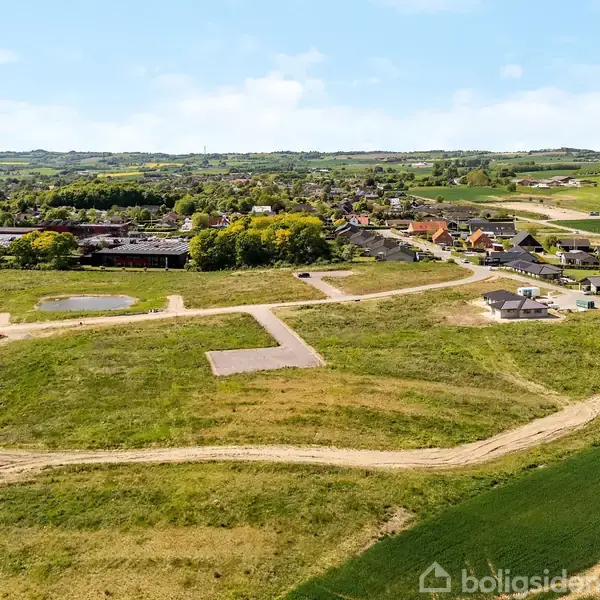 Luftfoto viser en åben byggegrund med tilkørselsveje omgivet af marker og bebyggelse i baggrunden. Landskabet er grønt med klare veje og spredte huse, perspektiv over kuperet terræn.