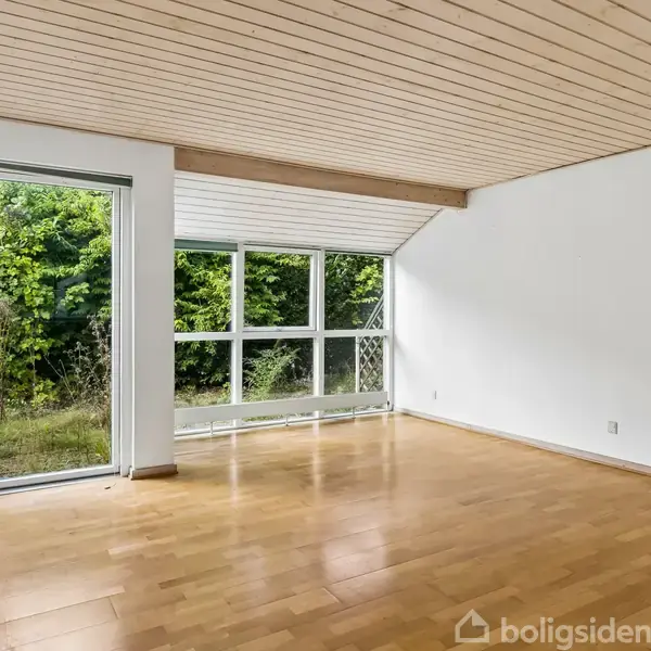 Et lyst værelse med trægulv og træbeklædt loft. Store vinduer og en dør åbner ud mod en frodig grøn have. Ingen tekst at transskribere.