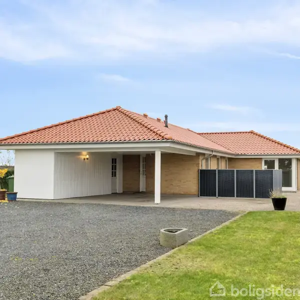 Huset står stille med rødt tegltag, flisebelagt carport, omgivet af grusopkørsel og velplejet græsplæne. Hækken skaber en grøn ramme under den lyseblå himmel.