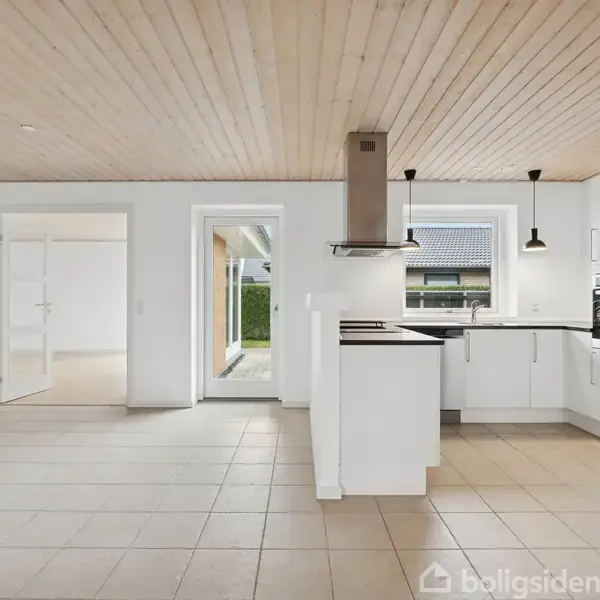 Et hvidt køkken med moderne indretning står tomt. Køkkenelementerne omfatter en ovn og kogeplade. Store vinduer giver udsigt til en have. Flisegulv og træloft præger rummet.