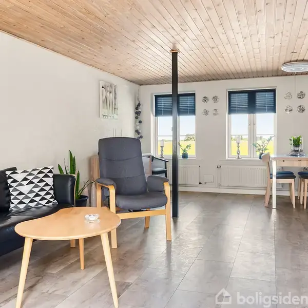 Sort sofa og lænestol står i en stue med trægulv, hvide vægge, træloft og runde sofabord. Tre vinduer med blå gardiner lukker lys ind. En spiseplads ses i baggrunden.