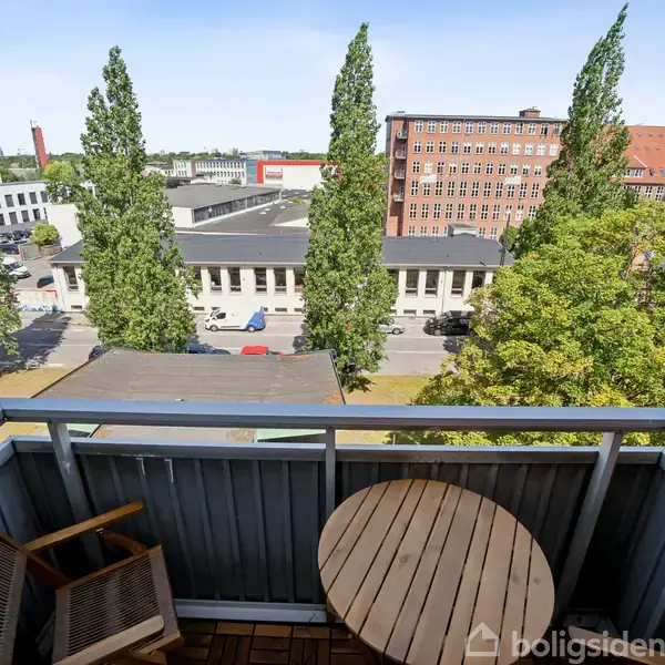 Balkon med rundt træbord og en stol. Udsigt over byområde med høje træer, en vej og røde murstensbygninger i baggrunden.