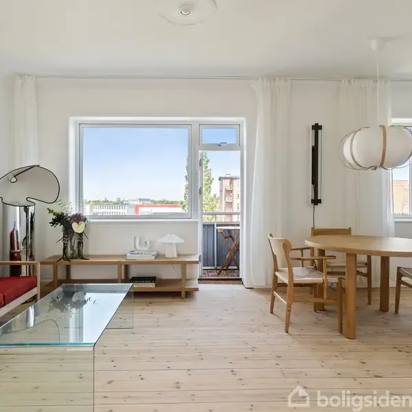Stue med rød sofa og glasbord; ved siden af står et rundt spisebord med stole. Store vinduer giver lys og udsigt til altan. En moderne lampe hænger over bordet.