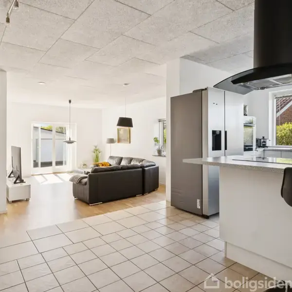 Køkken alrum med sort lædersofa, sofabord og fjernsyn i baggrunden. Et stort køkken-ø med høj stol og emhætte, moderne køleskab, og vinduer med haverudsigt.
