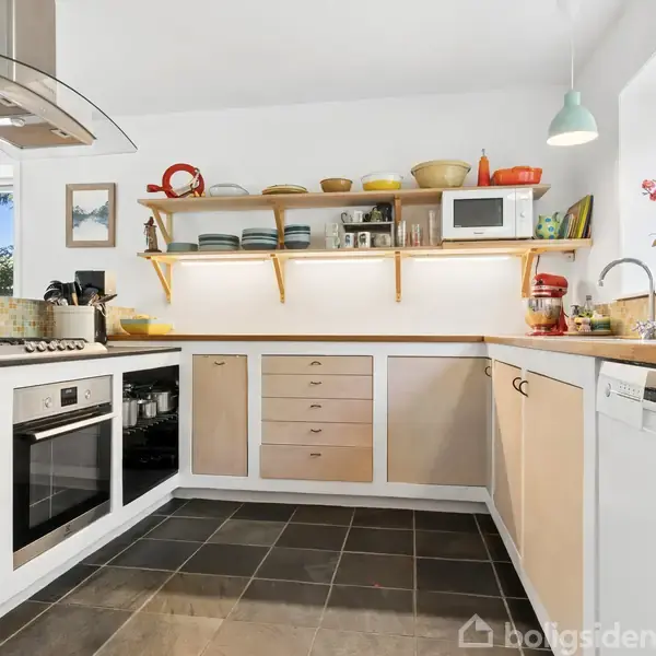 Et køkken med moderne indretning, hvor komfuret og ovnen er indbygget i en beige skabsenhed. Ovn, hylder med service, og vinduer lader dagslys strømme ind i rummet.
