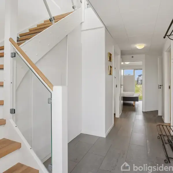 Trappe med trætrin og glasgelænder fører opad i en moderne, lys entré. Væggene er hvide, og gulvet belagt med store grå fliser. En skohylde og knagerække er til højre.