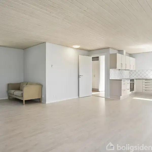 En lys stue med trægulv og et åbent køkken. En beige sofa står nær et vindue, der giver udsigt til haven. Køkkenet har hvide skabe og flisebelægning.