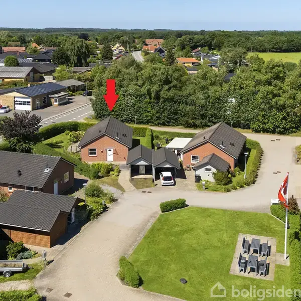 En rød pil peger på et rødt murstenshus med mørkt tag i en lille boligklynge omgivet af grønne træer og marker. I forgrunden ses en flagstang og en have.
