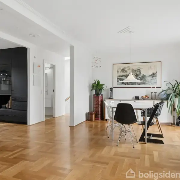 Spisestue med rundt bord og fire stole på trægulv i lys stue. Sort køkken baggrund. Grønne planter og kunst hænger på væggen. Moderne, minimalistisk indretning.