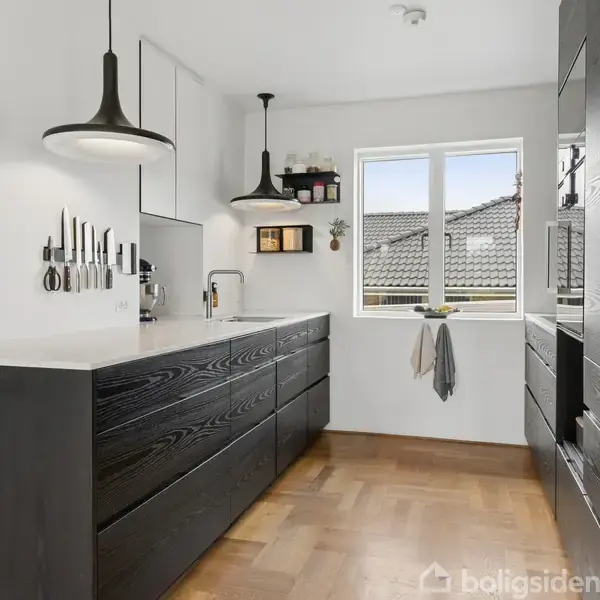 Sort køkkenbord med integreret vask og knive på væggen. Lamper hænger over bordet. Et vindue i midten giver udsigt til taget. Parketgulv og lyst interiør.