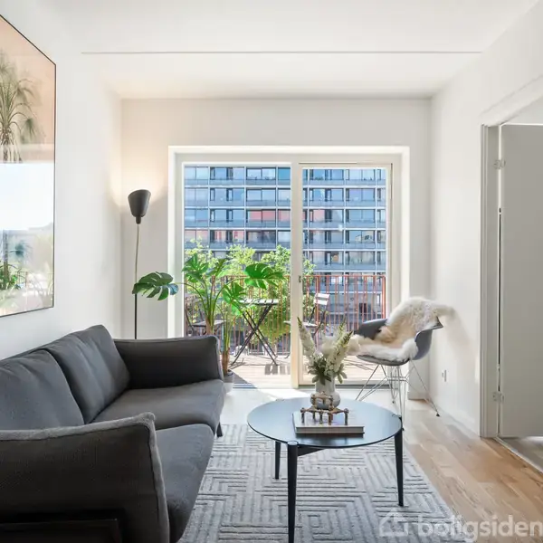 En grå sofa står i en moderne stue, med et rundt sofabord foran. Store vinduer i baggrunden åbner ud mod en altan med planter, omgivet af flere etagebygninger.