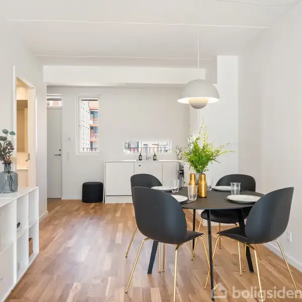 Spisebord med fire sorte stole står i en moderne spisestue med lyse trægulve og kunstnerisk pynt. En stor lampe hænger over bordet, og der er grønne planter i baggrunden.