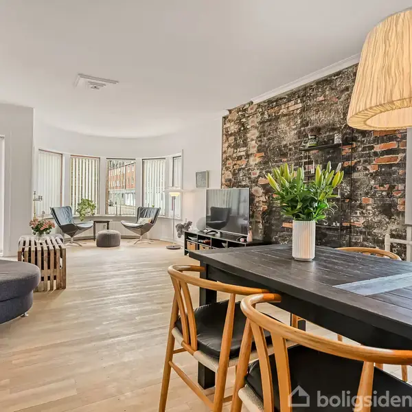Spisebord med træstole står i fokus under en ophængt lampe, mens en stue med sofa, lænestole og en rå murstensvæg skaber en hyggelig atmosfære i baggrunden.