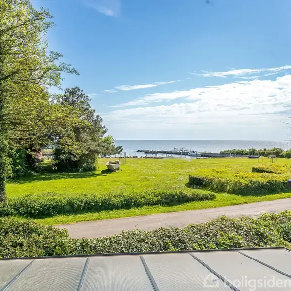 Et åbent vindue indrammer en udsigt over en grøn græsplæne med træer til venstre og en vej. I baggrunden ses en stille kystlinje med havet og en bådbro.