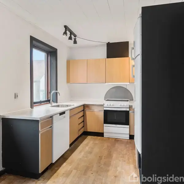 Køkkenet har lyse træskabe og en ovn under en emhætte. En håndvask er placeret ved vinduet. Rummet har trægulv og hvidmalede vægge med sort indramning.