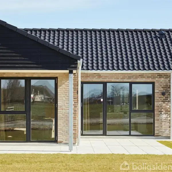 En moderne murstensvilla står med store, sorte vinduer, som reflekterer et grønt græsareal. Taget er mørkt og flisebelagt foran huset, der ligger i et roligt boligområde.