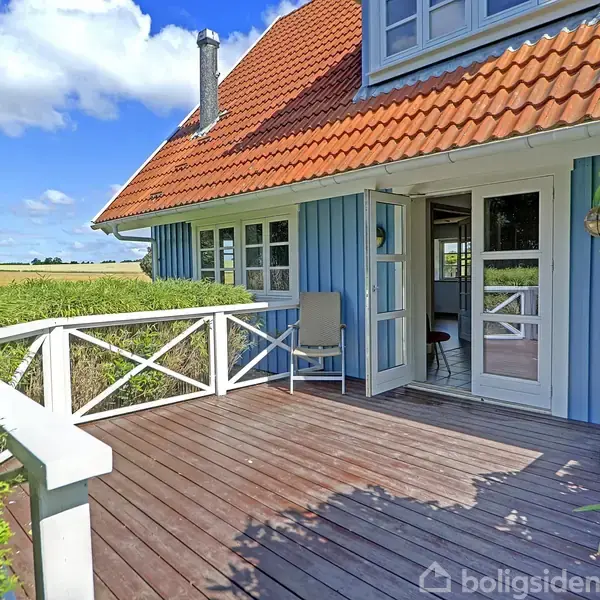 Blåt træhus med rødt tegltag har en åben terrassedør ud mod en solrig træterrasse. Omgivet af frodige marker og under en klar blå himmel med spredte skyer.